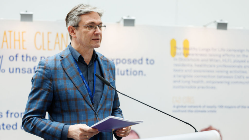 Adam Jarubas MEP, Chair of the SANT Subcommittee, speaking during healthy lungs for life exhibition at the European Parliament. He is wearing a blue suit with glasses and is stood behind a lectern. Behind him are boards with information about lung disease in Europe.