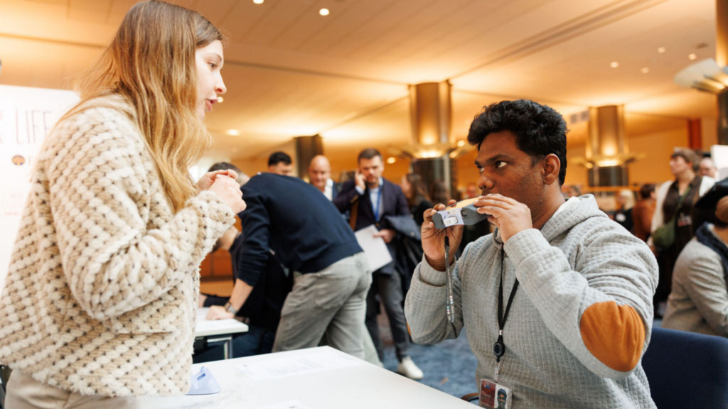 Two people are photographed at the European Parliament during healthy lungs for life exhibition. On the right is a volunteer tester helping the other person in the photo to complete a spirometry test. The person being tested is blowing into a spirometer to measure how much air they can blow out of their lungs.