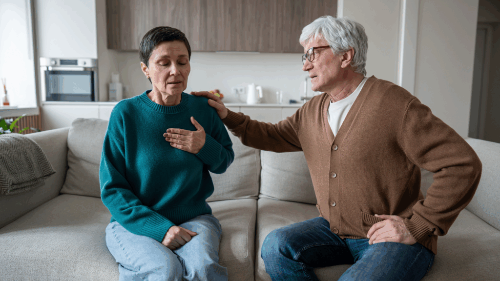 A lady holds her hand to her chest and sits on the sofa next to a man who comforts her.
